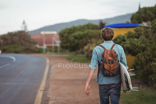 Visão traseira do homem com mochila carregando uma prancha de surf andando ao longo da estrada — Fotografia de Stock