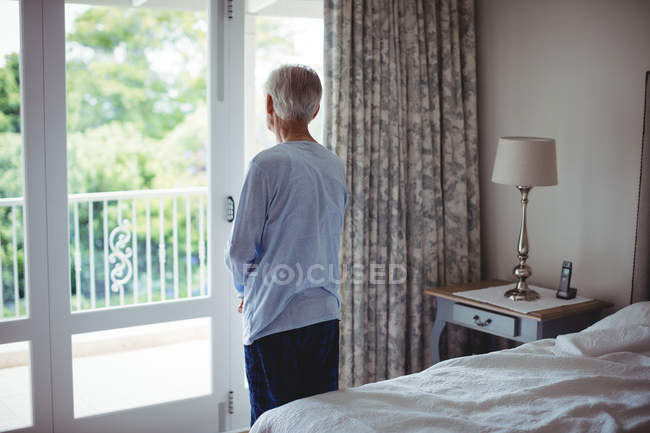 Homme âgé regardant par la fenêtre dans la chambre à coucher à la maison — Photo de stock