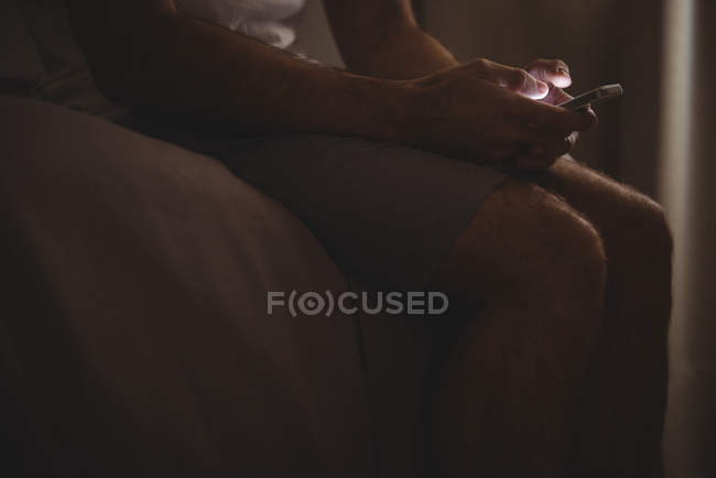 Средняя часть мужчины, сидящего на кровати в спальне, используя свой мобильный телефон — стоковое фото