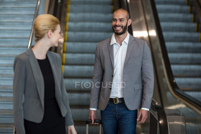 Gente de negocios sonriente con equipaje bajando en escaleras mecánicas en la terminal del aeropuerto - foto de stock