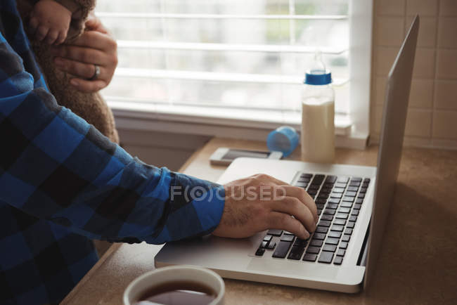 Manos del padre usando el ordenador portátil mientras sostiene al bebé en la cocina - foto de stock