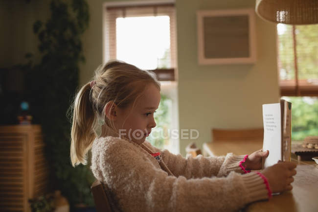 Ragazza bionda seduta a tavola e leggere libro a casa — Foto stock