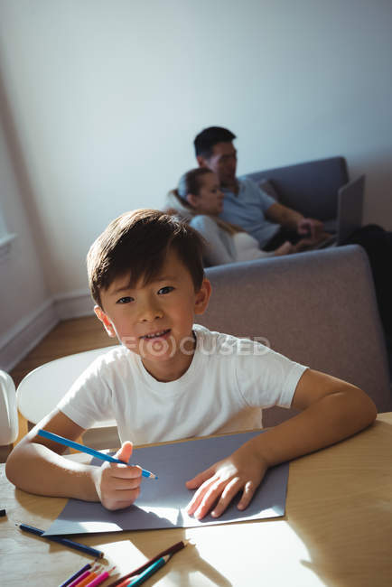 Retrato de un niño feliz dibujando en papel mientras los padres usan la computadora portátil en segundo plano - foto de stock