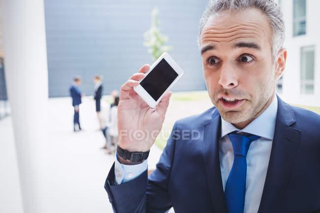 Businessman holding mobile phone and frowning outside office building — Stock Photo