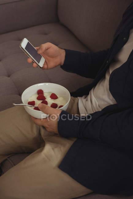 Partie médiane de l'homme utilisant le téléphone portable tout en prenant le petit déjeuner sur le canapé — Photo de stock
