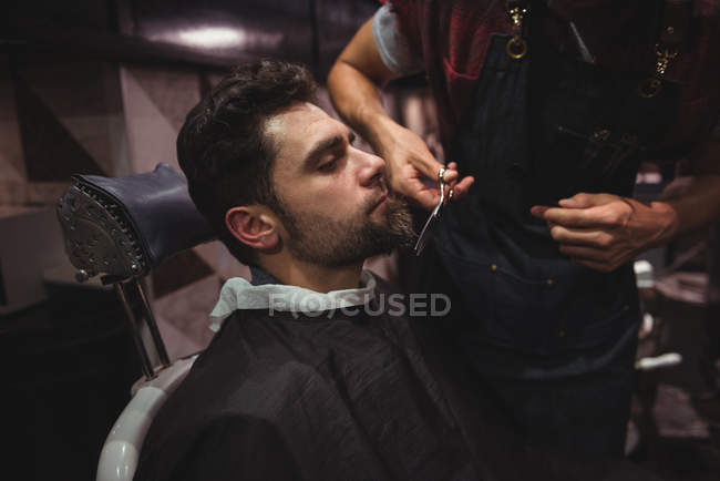 Мужчина стрижёт бороду парикмахером с ножницами в парикмахерской — стоковое фото