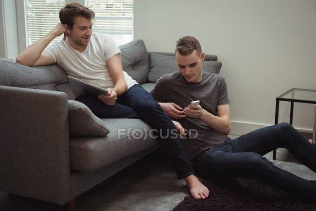Pareja gay usando tableta digital y teléfono móvil en la sala de estar en casa - foto de stock