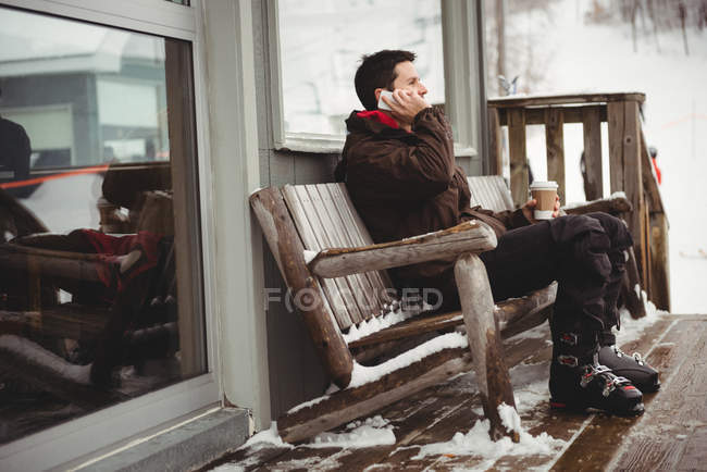 Hombre en el teléfono sentado en un banco en una estación de esquí disfrutando de la vista a la montaña - foto de stock