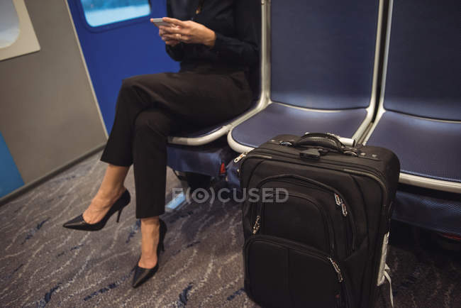 Sección baja de la mujer de negocios que usa el teléfono mientras está sentada con el equipaje en tren - foto de stock