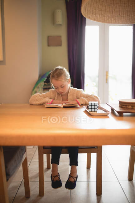 Linda chica sentada en la mesa y leyendo libro en casa - foto de stock