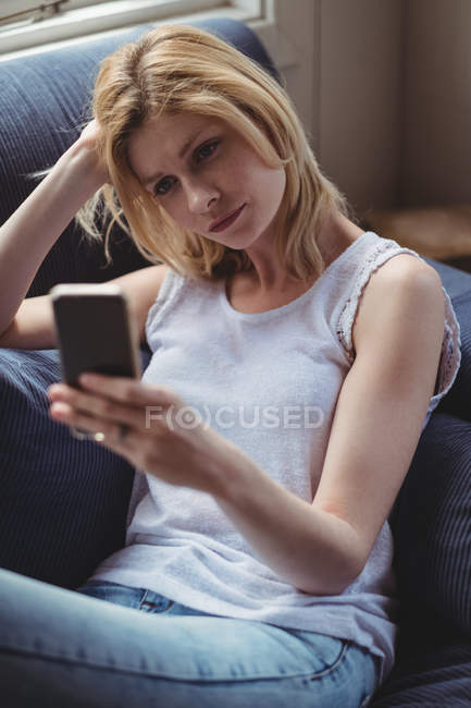 Mulher bonita sentada no sofá e usando telefone celular na sala de estar em casa — Fotografia de Stock