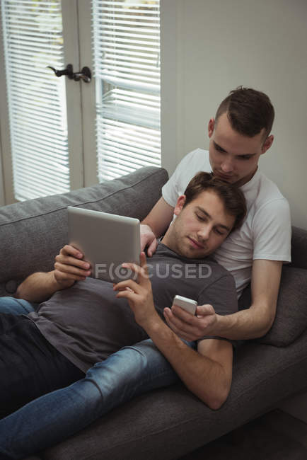 Schwules Paar benutzt digitales Tablet und Handy, während es zu Hause auf dem Sofa liegt — Stockfoto