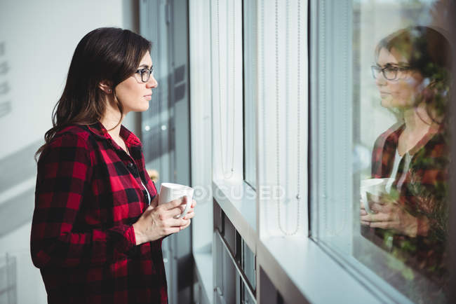 Задумчивая женщина смотрит в окно, выпивая кофе в офисе — стоковое фото