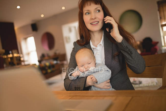 Mother talking on mobile phone while holding baby at home — Stock Photo
