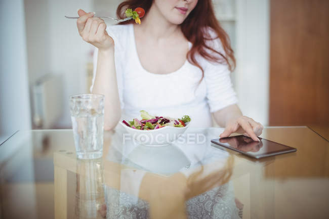 Вагітна жінка використовує цифровий планшет, маючи салат вдома — стокове фото