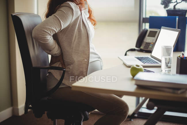 Pregnant businesswoman holding painful back while sitting on chair in office — Stock Photo