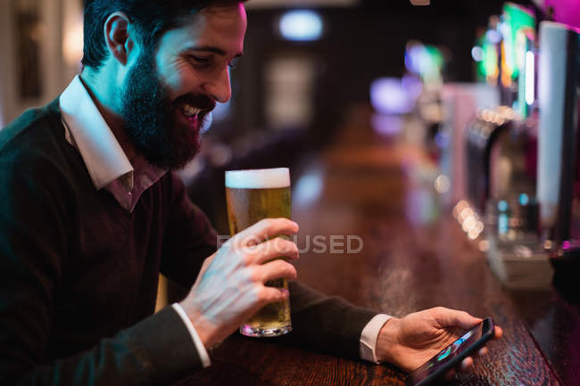 Hombre mirando el teléfono móvil mientras toma un vaso de cerveza en el mostrador del bar - foto de stock