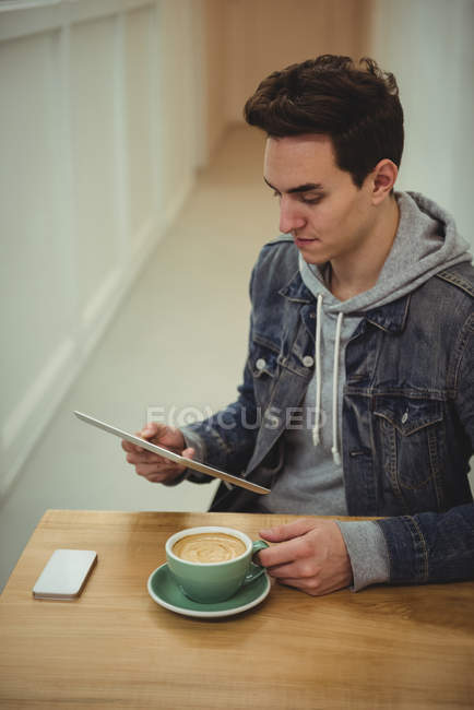 Чоловік використовує цифровий планшет, тримаючи чашку кави на дерев'яному столі в кав'ярні — стокове фото