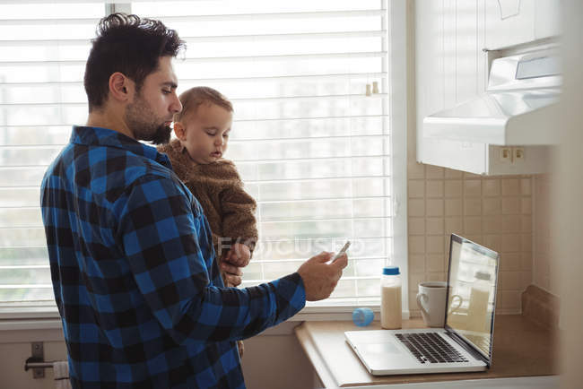 Отец использует мобильный телефон, держа ребенка на кухне дома — стоковое фото