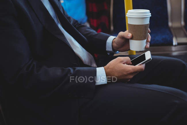 Sección media del hombre de negocios sosteniendo una taza de café desechable y utilizando el teléfono móvil en el autobús - foto de stock