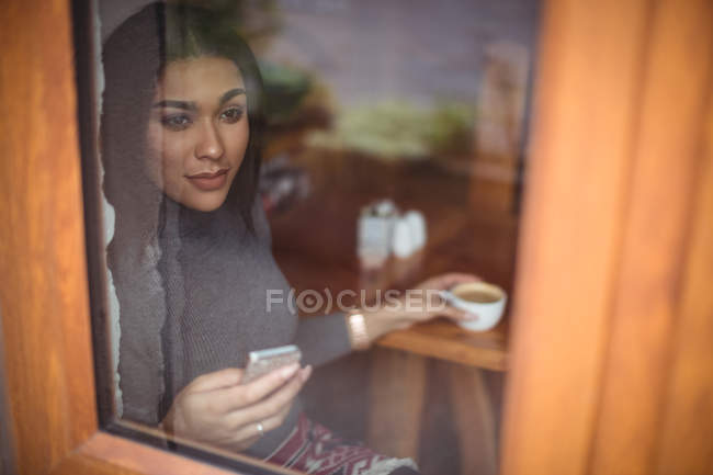 Mujer pensativa tomando una taza de café mientras usa el teléfono móvil en la cafetería - foto de stock