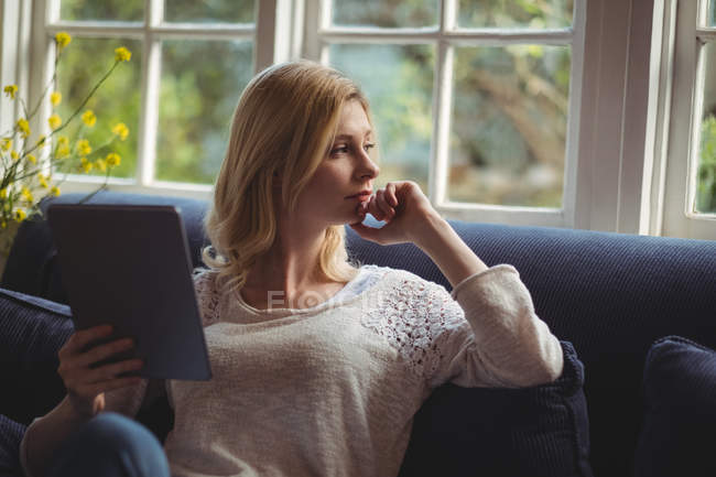 Задумчивая женщина сидит на диване с цифровым столом в гостиной на дому — стоковое фото