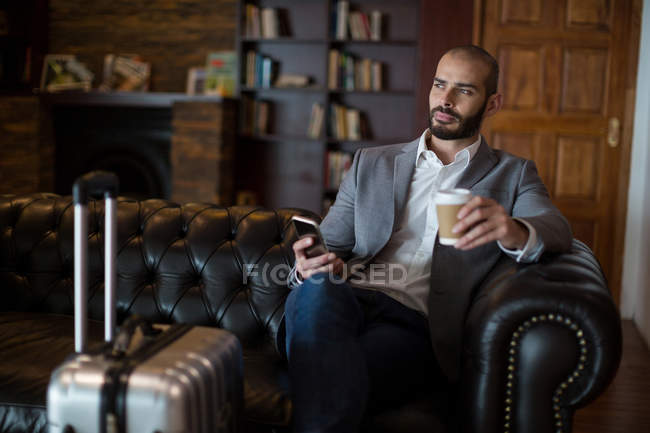 Thoughtful businessman holding mobile phone and coffee cup in waiting area at airport terminal — Stock Photo