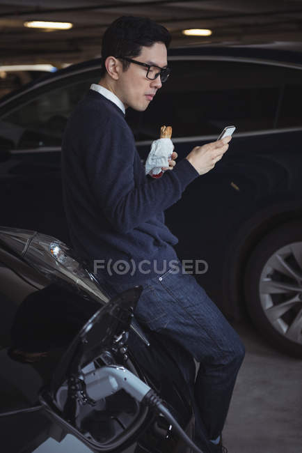 Man using mobile phone and eating snack while charging electric car in garage — Stock Photo
