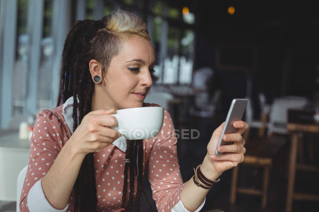 Mujer usando el teléfono móvil mientras toma café en la cafetería - foto de stock