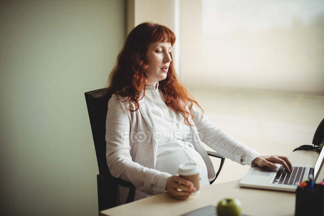 Femme d'affaires enceinte utilisant un ordinateur portable tout en prenant un café au bureau — Photo de stock