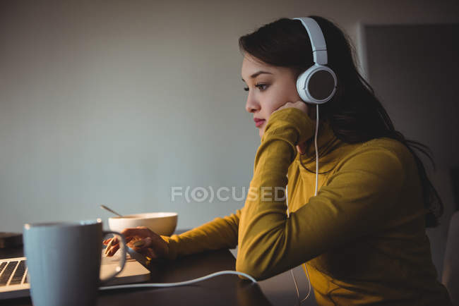 Mulher ouvindo fones de ouvido enquanto usa laptop na sala de estudo em casa — Fotografia de Stock