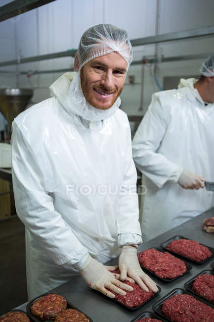 Retrato de carniceros sonrientes arreglando carne picada en bandeja de embalaje en fábrica de carne - foto de stock