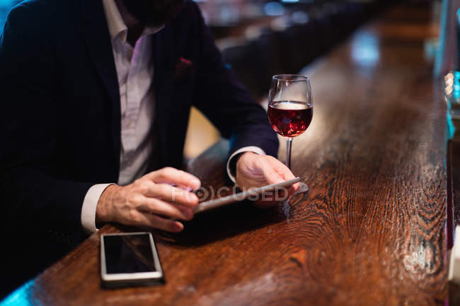 Empresário usando tablet digital com copo de vinho e telefone celular no balcão no bar — Fotografia de Stock