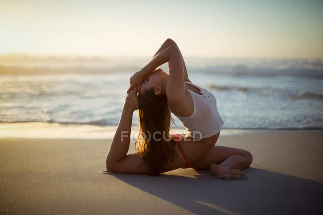 Mulher realizando exercício de alongamento na praia durante o pôr do sol — Fotografia de Stock