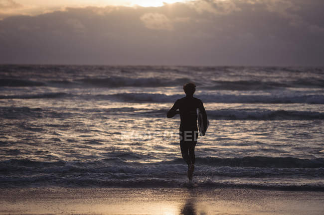 Силует чоловіка, що носить дошку для серфінгу, що біжить до моря в сутінках — стокове фото
