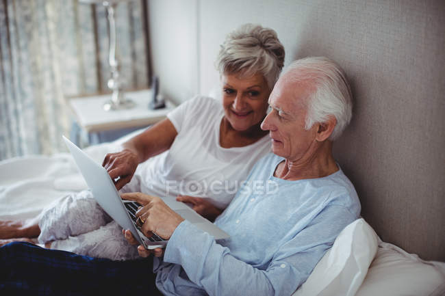 Пожилая пара с ноутбуком на кровати в спальне — стоковое фото