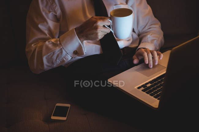 Средняя секция бизнесмена, использующего ноутбук за чашкой кофе дома — стоковое фото