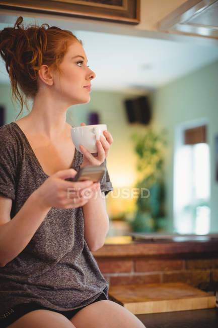 Продумана жінка використовує мобільний телефон, маючи каву вдома — стокове фото