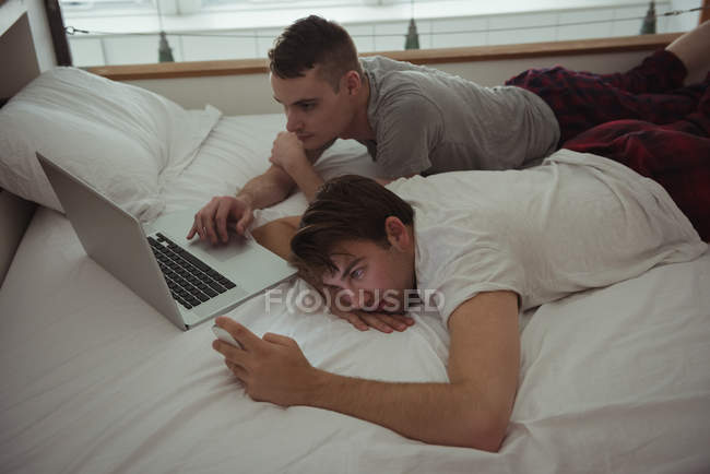 Couple gay utilisant téléphone mobile et ordinateur portable sur le lit dans la chambre — Photo de stock