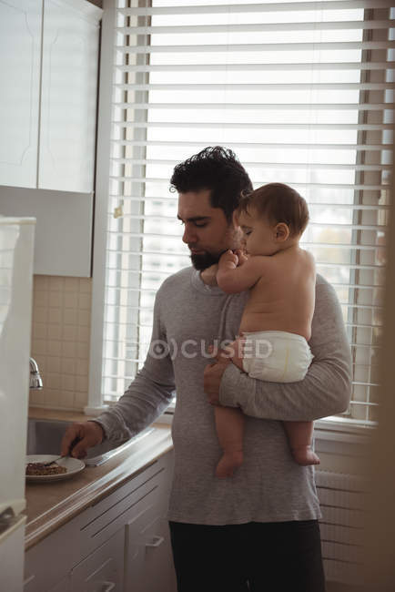 Padre preparando el desayuno mientras sostiene al bebé en la cocina - foto de stock