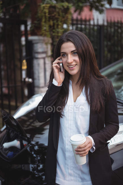 Woman talking on mobile phone while car being charged in background at electric vehicle charging station — Stock Photo