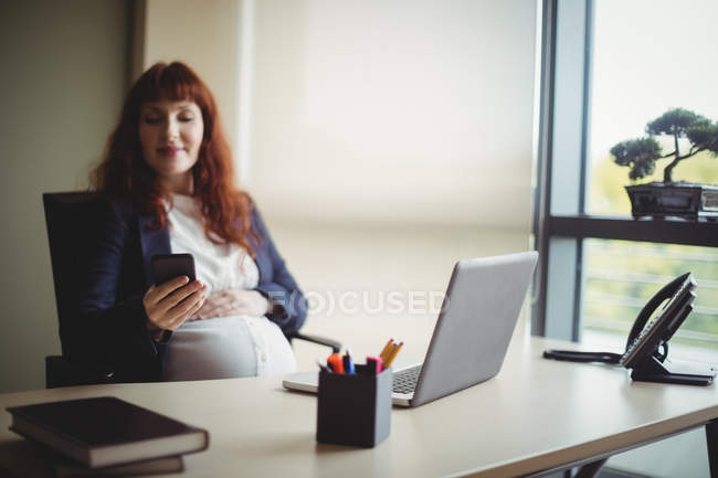 Беременная деловая женщина с помощью мобильного телефона в офисе — стоковое фото