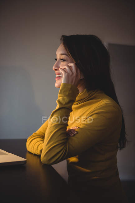Mujer hablando en el teléfono móvil mientras trabaja en el ordenador portátil en la sala de estudio - foto de stock
