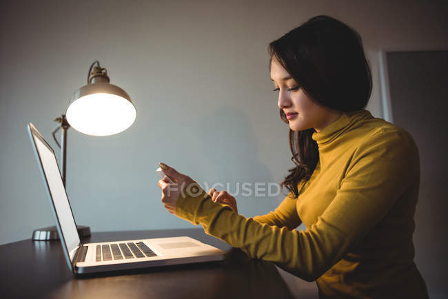 Жінка використовує мобільний телефон під час роботи на ноутбуці в навчальному залі вдома — стокове фото