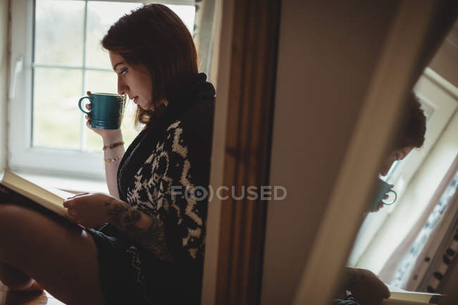 Mujer tomando café y leyendo un libro mientras está sentada en el alféizar de la ventana en casa - foto de stock