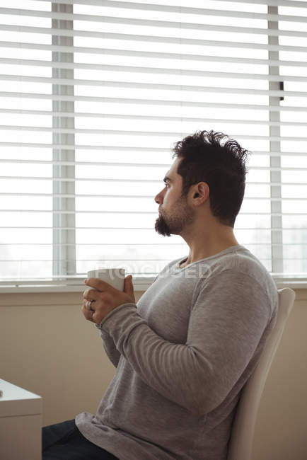 Homem pensativo segurando xícara de café em casa e olhando para longe — Fotografia de Stock
