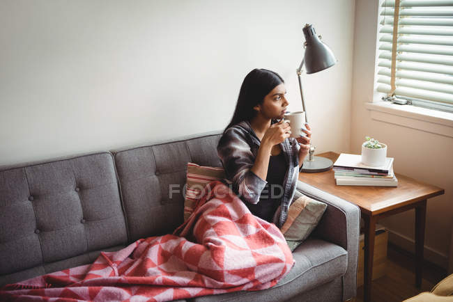 Donna che tiene la tazza di caffè guardando attraverso la finestra a casa — Foto stock