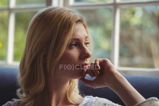Femme réfléchie regardant par la fenêtre dans le salon à la maison — Photo de stock