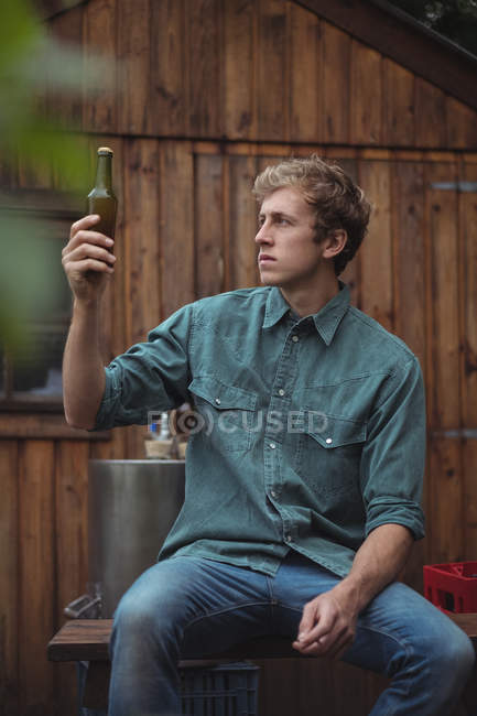 Чоловік сидить за межами домашньої пивоварні, дивлячись на пляшку пива — стокове фото