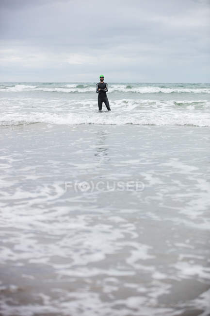 Athlete in wet suit standing with his hands crossed in the sea on beach — Stock Photo